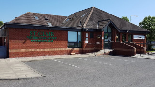 A brick veterinary clinic with sloped roofs and a ramp, surrounded by a parking lot and trees. Text on building:  A brick veterinary clinic with sloped roofs and a ramp, surrounded by a parking lot and trees. Text on building: