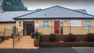 Building with beige walls and a gray roof, featuring a red door, surrounded by a black fence. Sign reads:  Building with beige walls and a gray roof, featuring a red door, surrounded by a black fence. Sign reads: