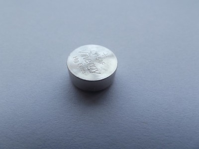 A small, cylindrical metal pill rests on a flat, gray surface. Engraved text reads: “H-F Ni MH very Hg free.”