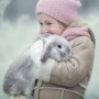 what-is-e-cuniculi-in-rabbits-vets-now A child in a pink beanie and scarf gently hugs a gray rabbit in a frosty, winter landscape with soft-focus, icy branches in the background.