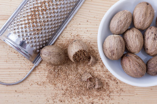 Whole nutmegs rest beside a metal grater on a wooden surface, with some ground nutmeg scattered around. A white bowl holds additional nutmegs nearby.