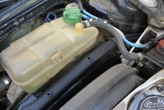 A translucent plastic coolant reservoir with a green cap is attached to hoses in a dusty car engine bay.