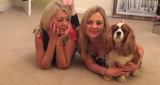 vets-now-pants-spaniel Two women and a Cavalier King Charles Spaniel lie on a carpeted floor, relaxed and smiling, with a framed forest picture leaning against the wall in the background.
