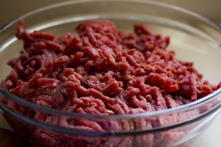 Ground beef sits in a clear glass bowl on a wooden surface, ready for preparation in a kitchen setting.