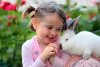 A young girl in pink, smiling warmly, gently embraces a white rabbit beside her; they are surrounded by vibrant green foliage and red flowers.