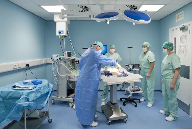 Medical team prepares in a hospital operating room, bright lights overhead, surrounded by medical equipment and monitors, wearing surgical gowns and masks, focused on a patient on a table.