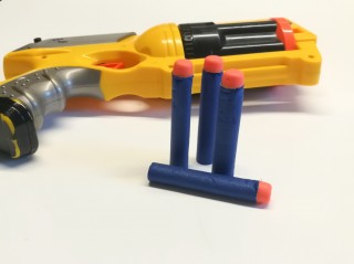Toy dart gun lies on a white surface, accompanied by three blue foam darts standing upright. The gun is yellow and silver with black accents, creating a playful scene.
