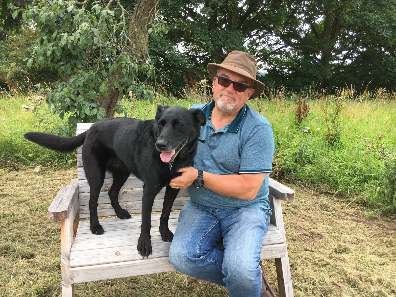 A man in a hat sits on a wooden bench holding a black dog panting, surrounded by grassy greenery and trees.