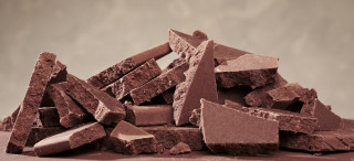 Rough chocolate chunks are stacked unevenly on a smooth surface, with a blurred, warm-toned background.