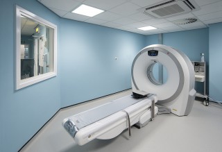 CT scanner rests idle in a clinical examination room, featuring light blue walls and overhead fluorescent lighting; a window overlooks a control room.