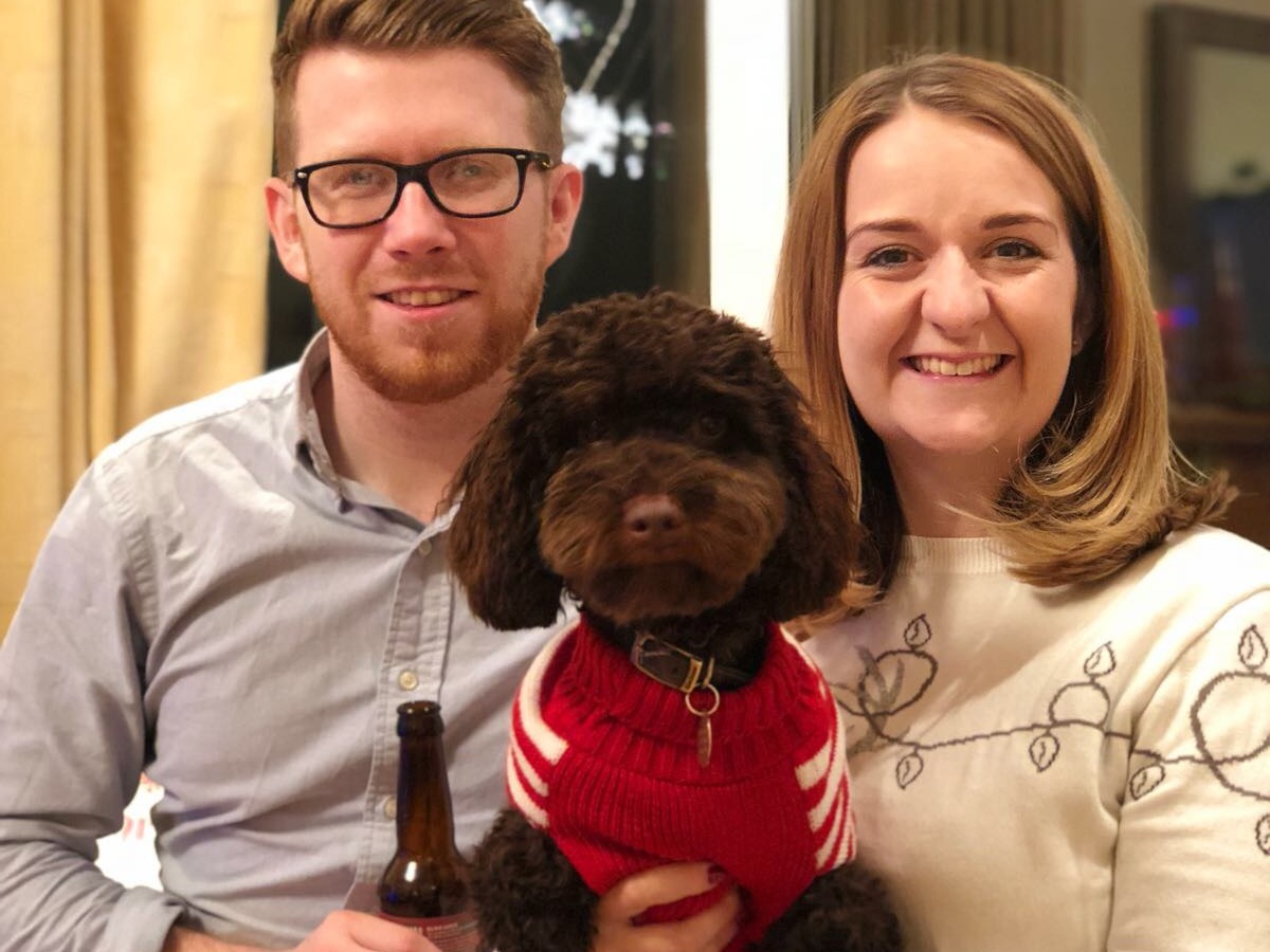A brown dog in a red sweater is held by two smiling people indoors, with one person holding a bottle.
