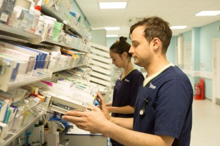 Two healthcare professionals organize medication shelves in a well-lit pharmacy. A man examines boxes labeled 