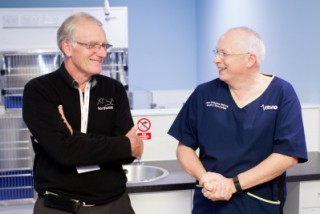 Two men stand in conversation, smiling, in a clinical setting. One wears a black sweater; the other, blue scrubs labeled 