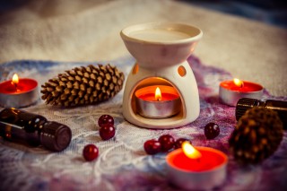 A lit tealight candle in a ceramic holder warms a room, surrounded by pinecones, essential oil bottles, and cranberries on a lace-patterned cloth.