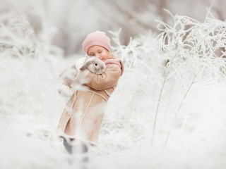 Child in a beige coat and pink hat hugs a fluffy rabbit in a snowy, frost-covered field.