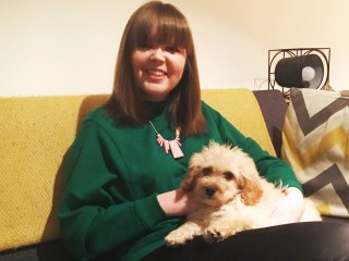 A woman wearing a green sweater sits on a yellow couch, smiling while holding a small, fluffy dog in her lap, beside patterned pillows and a decorative lamp.