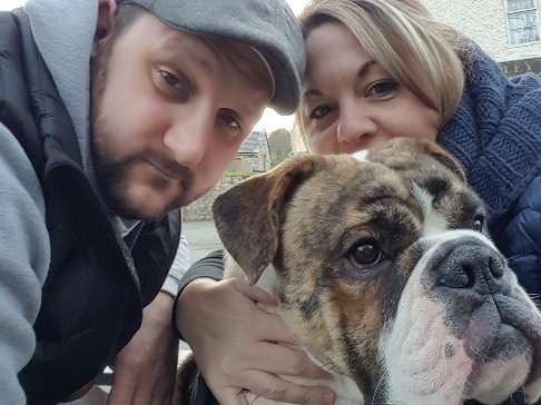 Two people crouch next to a brindle bulldog, gently holding it. They’re outdoors near a textured building with a window, appearing relaxed and content.