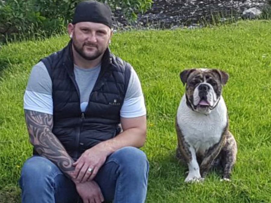 A man with a black cap and tattoo sits on grass beside a brindle bulldog. Behind them is a lush green area with a small stream.