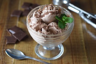 Chocolate ice cream sits in a glass bowl, garnished with chocolate shavings and a mint sprig, on a wooden table next to a spoon and square chocolate pieces.