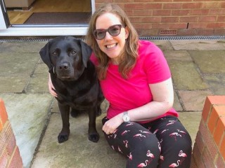 A woman in a pink shirt and flamingo-patterned pants smiles while sitting on a stone patio next to a black Labrador, with an open doorway in the background.