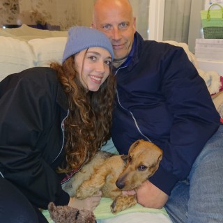 Dotty with her loving owners A dog lies between two people on a couch. The woman smiles, wearing a blue beanie, while the man in a dark jacket gently holds the dog. Cozy indoor setting.