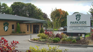 A veterinary clinic with a brick exterior features green signage and a logo. A person enters the building. The foreground has a garden with shrubs and a large sign:  A veterinary clinic with a brick exterior features green signage and a logo. A person enters the building. The foreground has a garden with shrubs and a large sign: