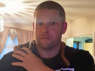 A man holds a snake draped over his shoulder in a dimly-lit room with curtains and soft lighting from a ceiling fixture.