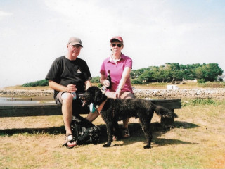 Two people sit on a bench holding bottles, with a large black dog standing beside them, in a grassy area near a rocky shoreline under a clear sky.