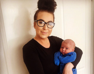 A woman wearing glasses and a black shirt holds a baby dressed in a blue onesie. They stand in front of a white door, smiling softly at the camera.