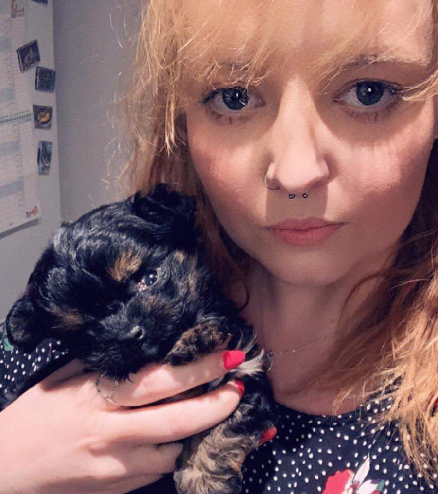 A young woman with a nose ring holds a small black and brown puppy in her arms, standing in a room with a calendar on the wall.
