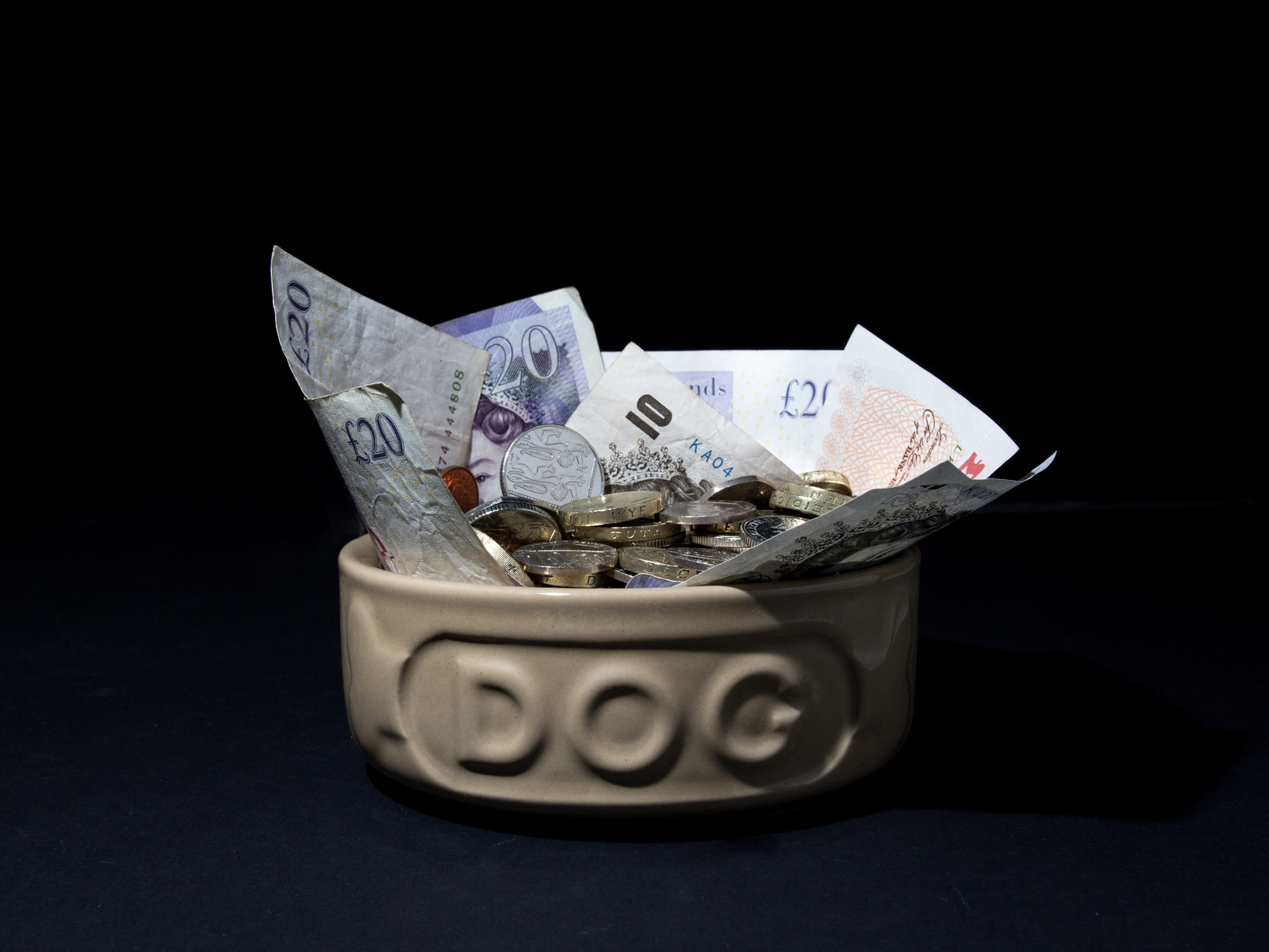 A dog bowl filled with British pound notes and coins, prominently marked with “DOG,” situated against a dark background.