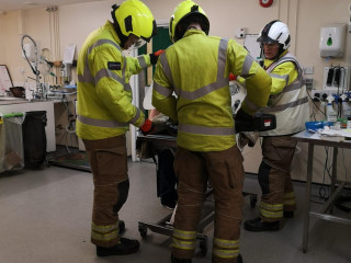 Three people in bright safety gear and helmets examine equipment in what appears to be a medical or laboratory setting, surrounded by tables, sinks, and various supplies.