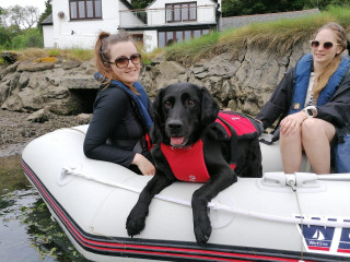 A black Labrador in a red harness lies on an inflatable boat with two women wearing life jackets, near a rocky shoreline and a white house.