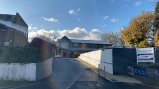 A building with a gabled roof stands at the end of a sloped driveway, flanked by brick and concrete structures, under a blue sky with clouds. Signs read  A building with a gabled roof stands at the end of a sloped driveway, flanked by brick and concrete structures, under a blue sky with clouds. Signs read
