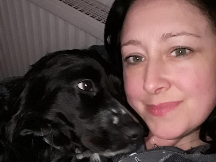 A black dog rests its head against a woman's face, both appearing relaxed and close, in a dimly lit indoor setting.