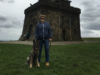 A person wearing sunglasses stands with a German Shepherd on a grassy hill in front of a stone building under a cloudy sky. Other people are seen in the background.