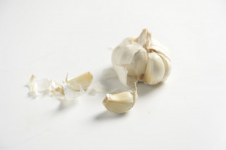 A garlic bulb with several peeled cloves rests on a white surface, surrounded by scattered papery skins.