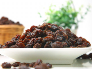 A pile of dried raisins sits on a white plate, surrounded by scattered raisins, with a blurred plant and basket in the background.