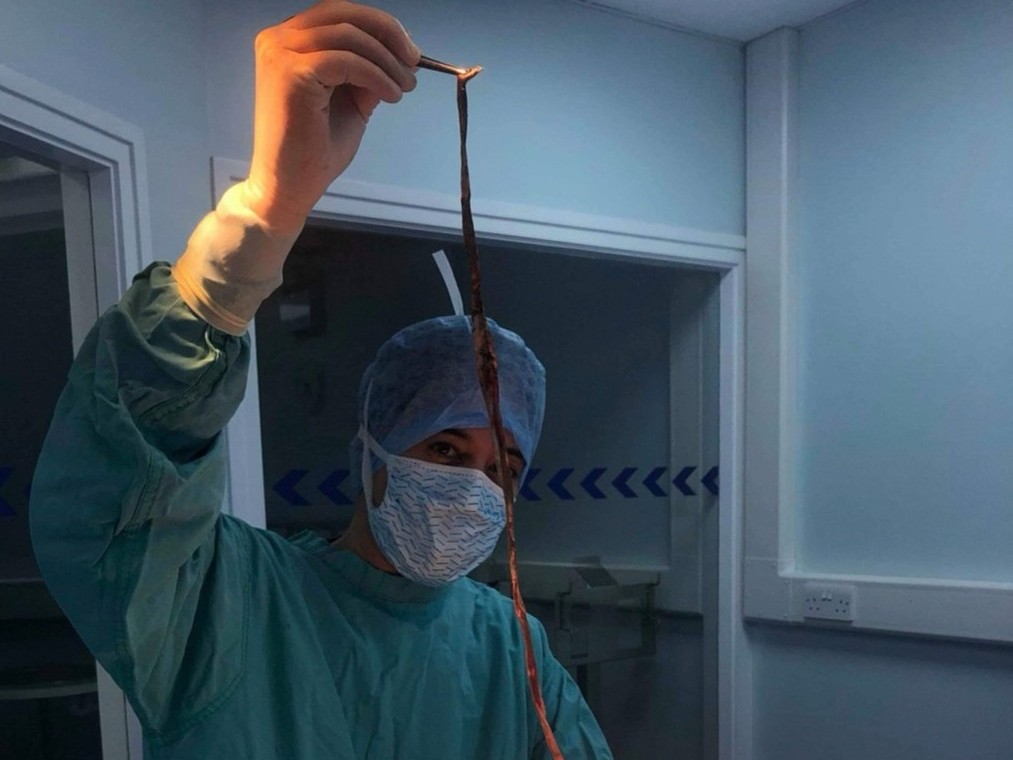 A medical professional in surgical attire holds a long object with forceps, standing in a clinical room with blue walls and glass windows.