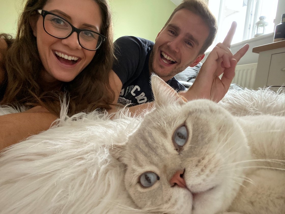 A fluffy white cat rests on a plush rug, while two smiling people lean beside it, indoors near a window. One person gestures with a peace sign.