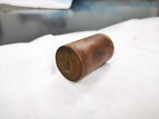 Karma's cork A cylindrical metal object rests on a white surface, displaying a rusty, textured appearance. The background is out of focus, suggesting an industrial or workshop setting.