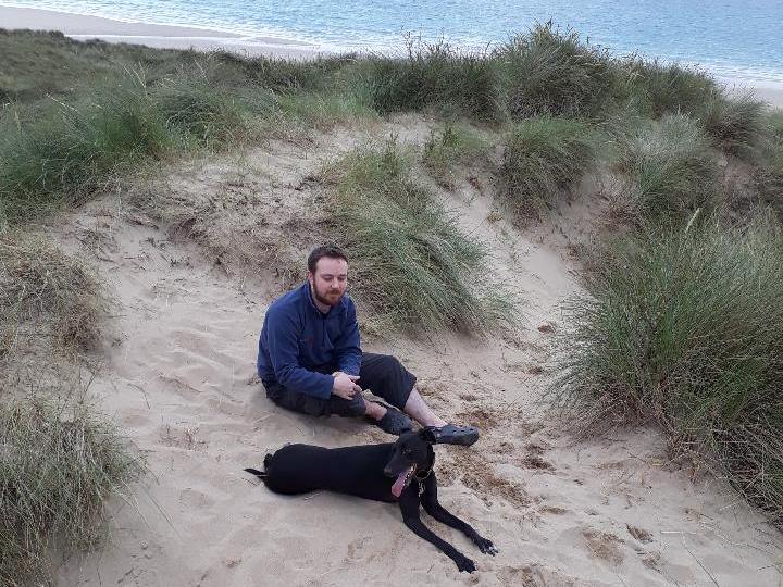 Sally the barbed wire dog A man sits beside a black dog on sandy dunes, surrounded by tall grass, with a beach and blue ocean in the background.