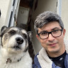 A dog with a gray and white coat sits beside a man wearing glasses, both outside a doorway. The dog's collar reads 