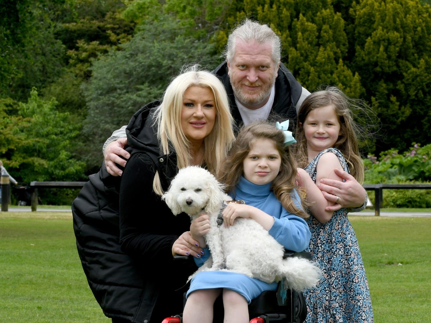A woman, man, and two girls, one holding a small white dog, pose closely together outdoors in a park, with green trees in the background.