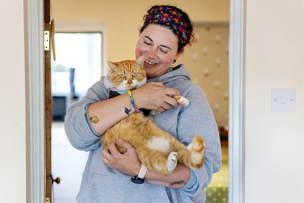 A person holds a relaxed orange cat in their arms, standing in a doorway. The room has light-colored walls and visible tattoos on the person's arms.
