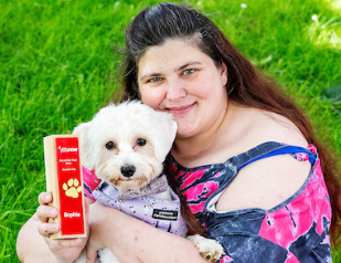 A woman holds a small white dog and a red award labeled 