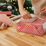 Woman wrapping a Christmas gift in red and white striped paper with gold ribbon on wooden table