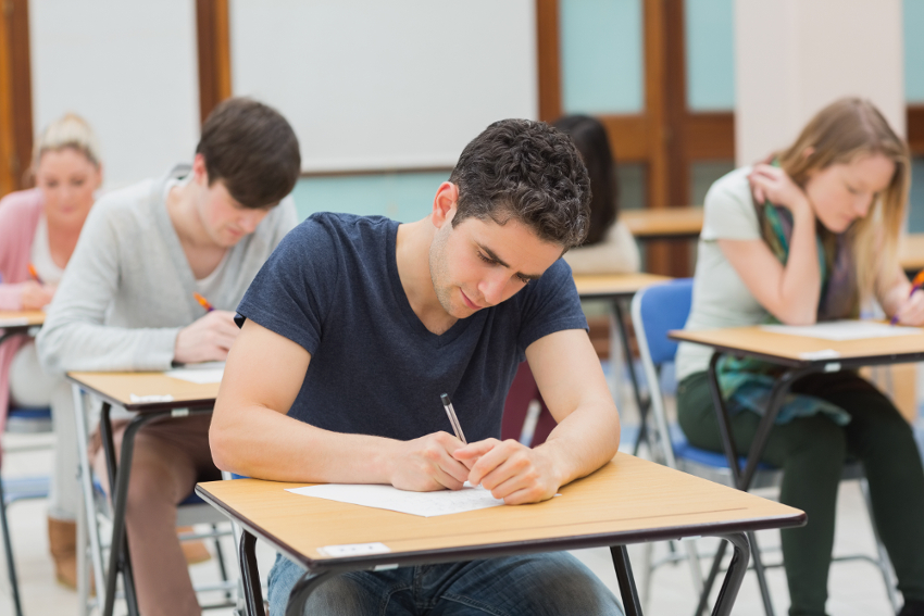 A group of students sits at individual desks, writing on papers in a classroom setting, focused and engaged in an exam or assignment.