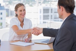 Two people, a woman and a man, shake hands across a table with a laptop and open notebook, in a bright office setting.