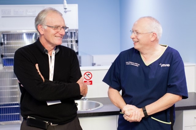 Two men are talking and smiling in a clinical setting. One wears a black sweater; the other is in blue scrubs with text: John Chitty BVetMed CertZooMed MRCVS ANZCVS.